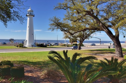 A white lighthouse stands by the shore, surrounded by greenery and a clear blue sky, with the ocean in the background.