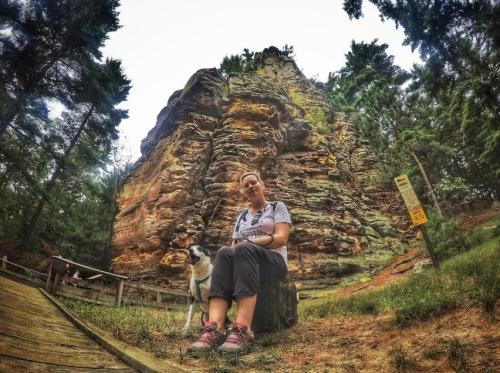 A woman sits on a log beside a dog, with a rocky cliff and trees in the background.