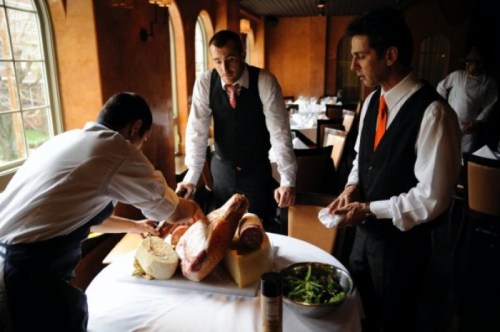 Three men in a restaurant setting examine a display of meats and cheeses on a table.