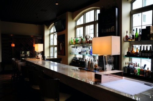 A sleek bar with a long countertop, illuminated lamps, and shelves stocked with various bottles in a stylish setting.