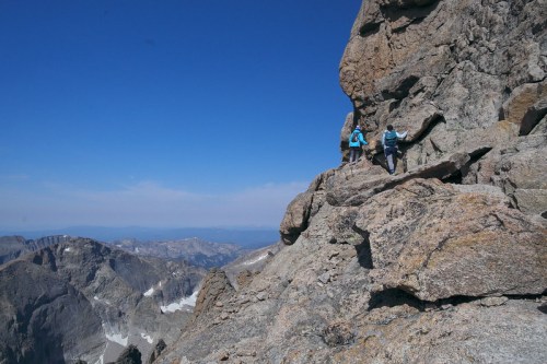 Two hikers navigate a rocky mountain path against a clear blue sky and distant mountain range.