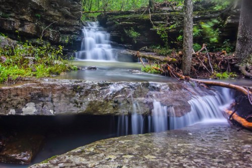 A serene waterfall cascades over rocky ledges, surrounded by lush greenery and trees in a tranquil forest setting.