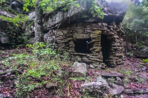 An old stone structure partially hidden by trees and foliage, surrounded by rocks and overgrown vegetation.