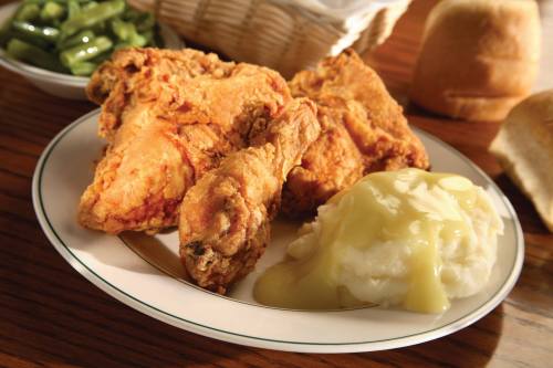 A plate of crispy fried chicken with mashed potatoes and gravy, served with green beans and a roll in the background.