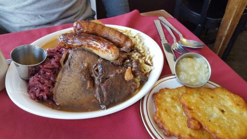 A plate of hearty food featuring beef brisket, sausages, red cabbage, potato pancakes, and applesauce on the side.