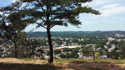 A scenic view from a hilltop overlooking a town, with trees and rolling hills in the background under a blue sky.
