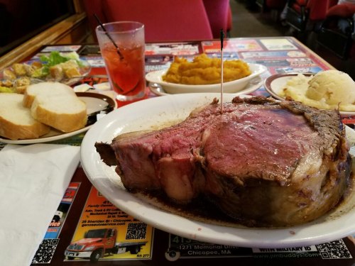 A large prime rib steak on a plate, accompanied by mashed potatoes, sweet potatoes, salad, bread, and a drink.