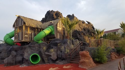 A tropical-themed water slide structure with green slides, surrounded by rocks and palm trees under a cloudy sky.