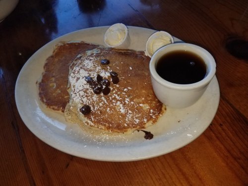 A plate of pancakes topped with powdered sugar and chocolate chips, served with butter and a small cup of syrup.