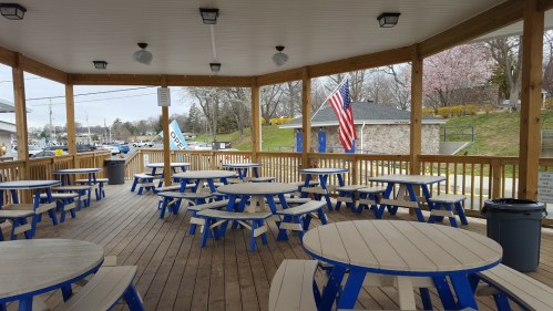 Outdoor seating area with blue and gray picnic tables under a wooden pavilion, featuring an American flag in the background.