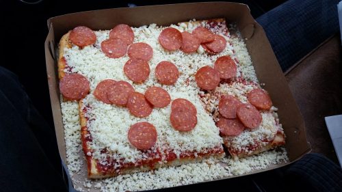 A rectangular pizza with a thick crust, topped with cheese and pepperoni slices, in a cardboard box.