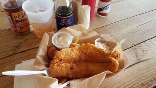 A plate of fried fish with fries, coleslaw, and dipping sauces, accompanied by drinks on a wooden table.