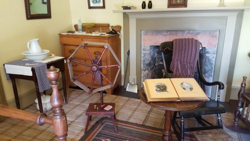 A vintage room featuring a spinning wheel, wooden furniture, a rocking chair, and historical photographs on a table.