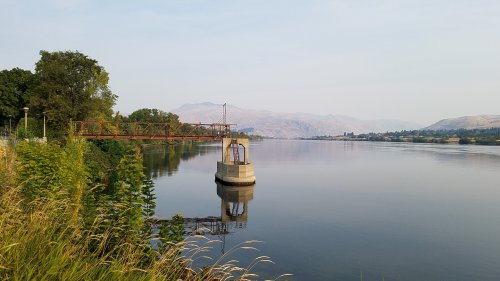 A calm river scene with a bridge, grassy banks, and mountains in the background under a clear sky.
