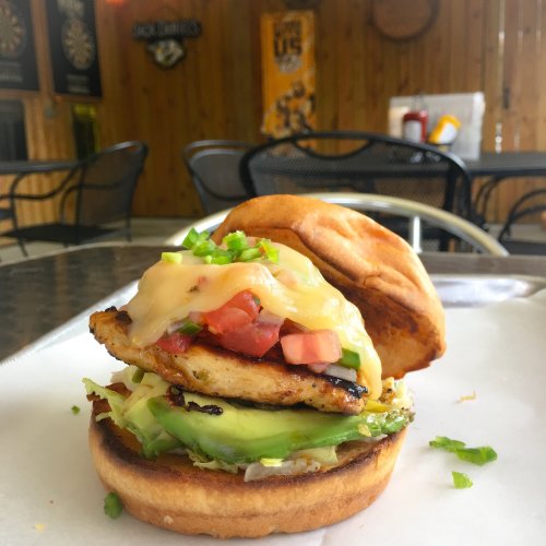 A grilled chicken sandwich with melted cheese, tomatoes, avocado, and green peppers on a toasted bun, served in a casual setting.