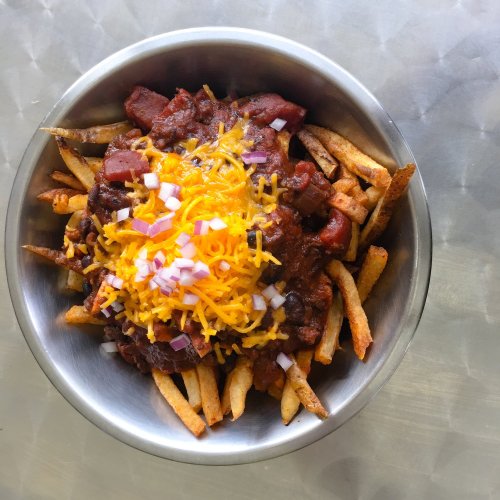 A bowl of fries topped with chili, shredded cheese, and diced onions on a metallic surface.