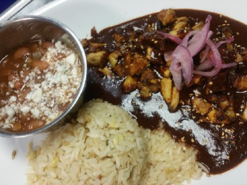 A plate featuring rice, beans in a bowl, and a serving of chicken with mole sauce, topped with pickled onions.
