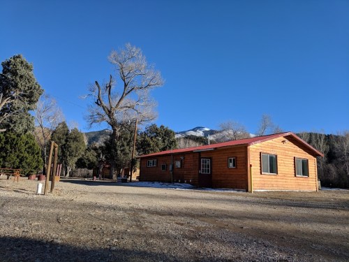 A wooden cabin with a red roof surrounded by trees and mountains under a clear blue sky.