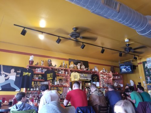 A lively restaurant interior with yellow walls, a bar, patrons seated, and various decorations on shelves.