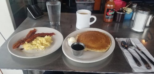 A breakfast plate with scrambled eggs, bacon, pancakes, syrup, and a cup of coffee on a metal table.