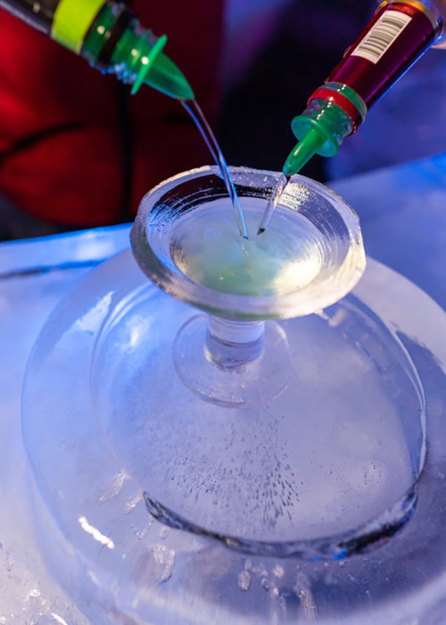 Two colorful liquid streams are being poured into a glass on an ice surface, creating a vibrant drink.