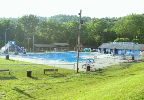 A sunny view of a municipal pool with slides, diving boards, and grassy areas, surrounded by trees and benches.