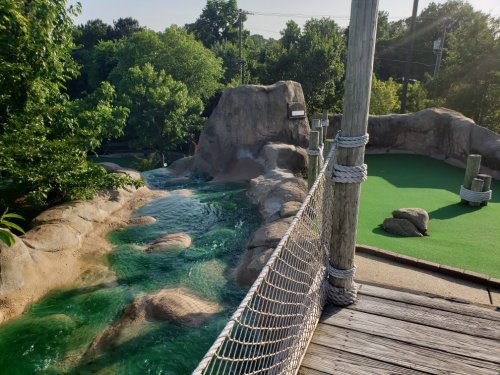 A scenic view of a mini-golf course featuring a rocky landscape, a small stream, and lush greenery.