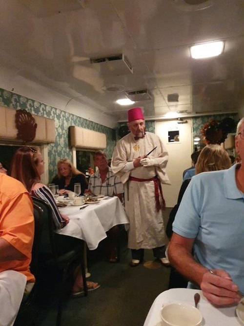 A man in traditional attire and a pink hat stands in a dining area, engaging with guests at tables.
