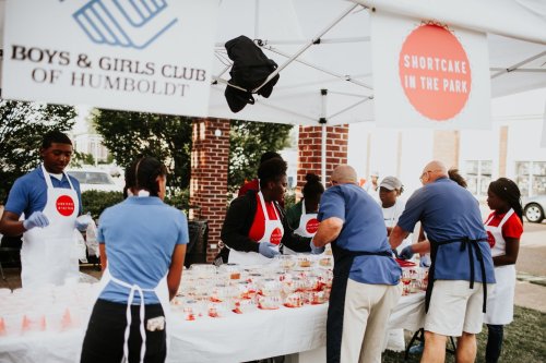 A group of people serving shortcake at a community event under a tent, with a Boys & Girls Club banner in the background.