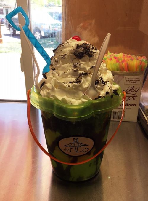 A colorful ice cream sundae in a plastic bucket, topped with whipped cream, crushed cookies, and a cherry.
