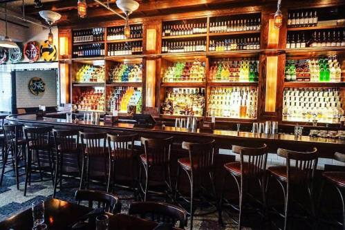 A well-stocked bar with colorful bottles on shelves, wooden counters, and empty stools in a cozy setting.