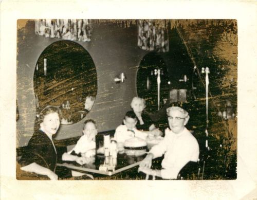 A vintage photo of a family at a restaurant, smiling around a table with circular mirrors in the background.