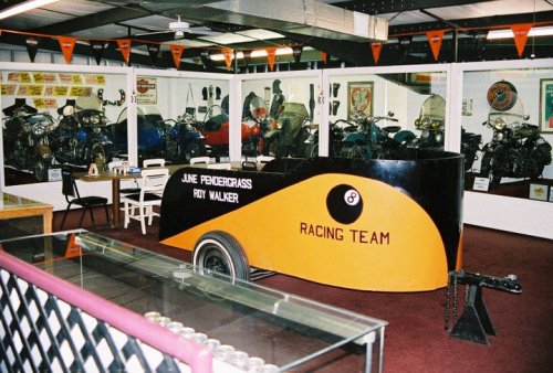 A vintage racing team display featuring a black and orange racing cart in a museum filled with motorcycles.