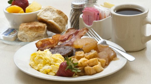 A breakfast plate with scrambled eggs, bacon, fruit, biscuits, and coffee on a table.