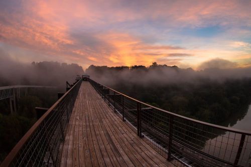 A wooden bridge stretches over a foggy landscape at sunrise, with vibrant colors in the sky and trees in the background.