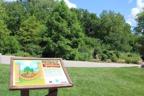A sign welcomes visitors to the Rain Garden, surrounded by lush greenery and trees under a blue sky.