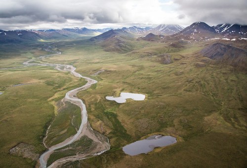 Aerial view of a winding river through a vast green landscape with mountains and small lakes under a cloudy sky.