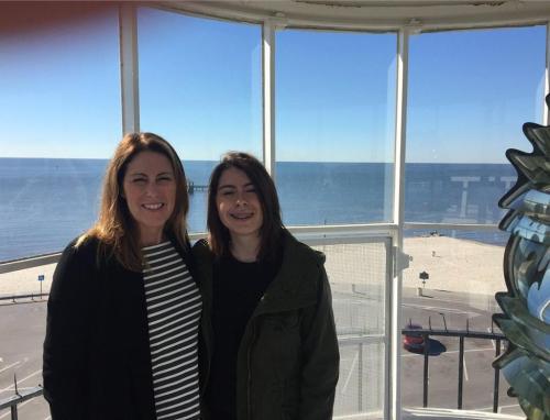Two women stand inside a lighthouse, smiling with a view of the beach and ocean in the background.