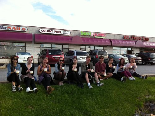 A group of ten people sitting on grass outside a store, each holding a cup, smiling and enjoying their time together.