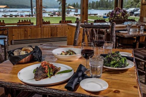 A rustic dining table with gourmet dishes, glasses of water and wine, overlooking a scenic lake view.