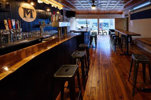 Interior of a bar featuring a wooden counter, stools, and a row of beer taps against a dark wall.