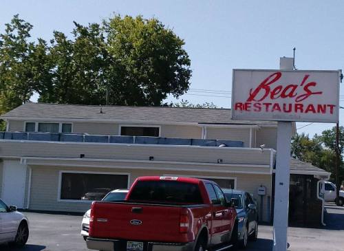 Sign for Bea's Restaurant in front of a building with parked cars and trees in the background.