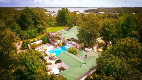 Aerial view of a resort with a pool, surrounded by trees and a lake in the background on a sunny day.