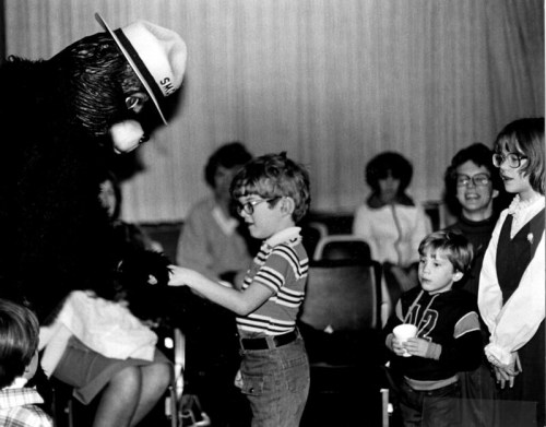 A child interacts with a costumed character while other children and adults watch in a seated audience.