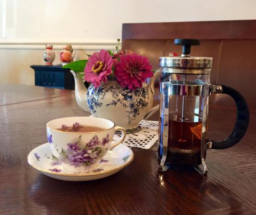A floral teacup and a French press sit on a wooden table, alongside a teapot and vibrant flowers.