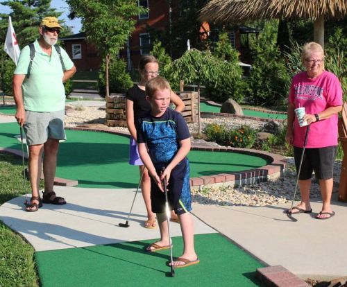 A group of four people enjoying mini-golf on a sunny day, with one child preparing to take a shot.
