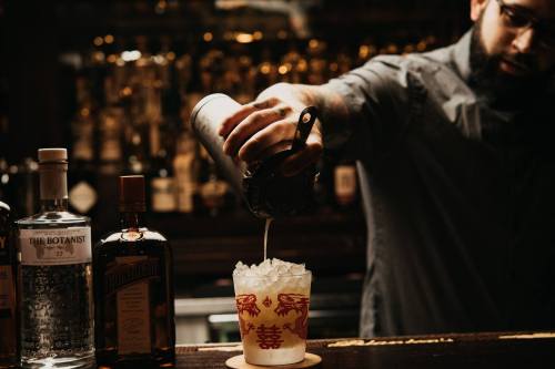 A bartender pours a cocktail over ice into a glass, with various bottles of spirits in the background.