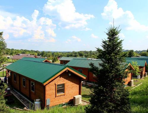 A scenic view of wooden cabins with green roofs, surrounded by lush greenery and a blue sky with fluffy clouds.