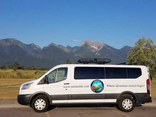 A white shuttle van parked with mountains in the background, featuring a logo that reads "Where adventure stories begin!"