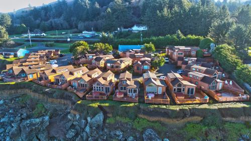 Aerial view of a coastal village with wooden cabins on a cliff, surrounded by trees and a road in the background.